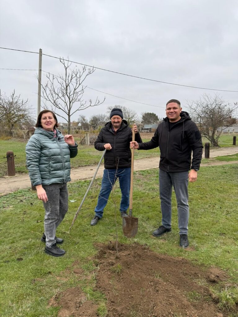 Троє дорослих на трав’янистій ділянці садять молоде дерево: один тримає лопату, інший — опору для саджанця, третій спостерігає. Земля вже розкопана, на тлі — голі дерева, дерев’яні стовпчики та хмарне небо. 