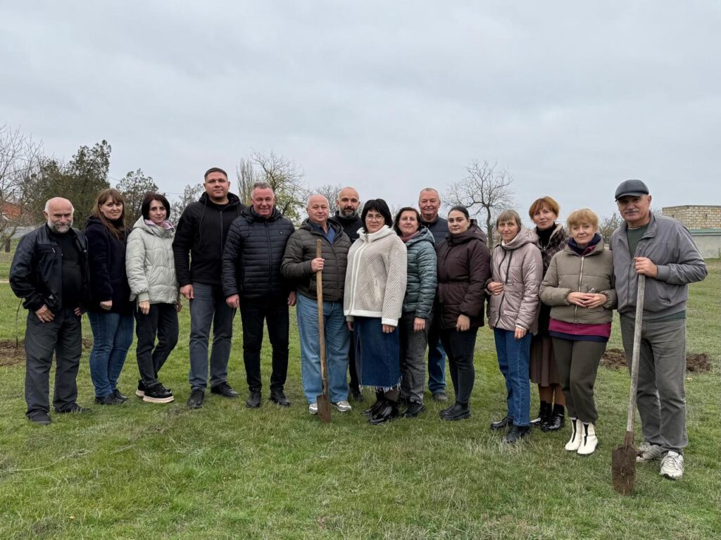 Шістнадцять дорослих стоять на трав’янистому полі в прохолодну пору року. Один із них тримає лопату — ймовірно, це спільна акція з озеленення чи догляду за територією. Усі вдягнені по-осінньому, на тлі — голі дерева та хмарне небо.