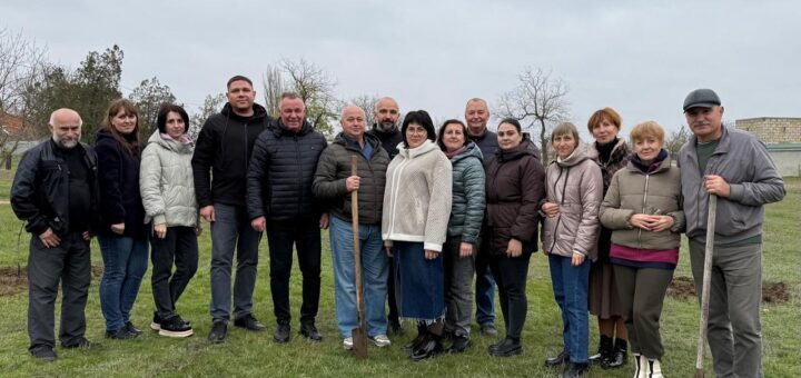 Шістнадцять дорослих стоять на трав’янистому полі в прохолодну пору року. Один із них тримає лопату — ймовірно, це спільна акція з озеленення чи догляду за територією. Усі вдягнені по-осінньому, на тлі — голі дерева та хмарне небо.