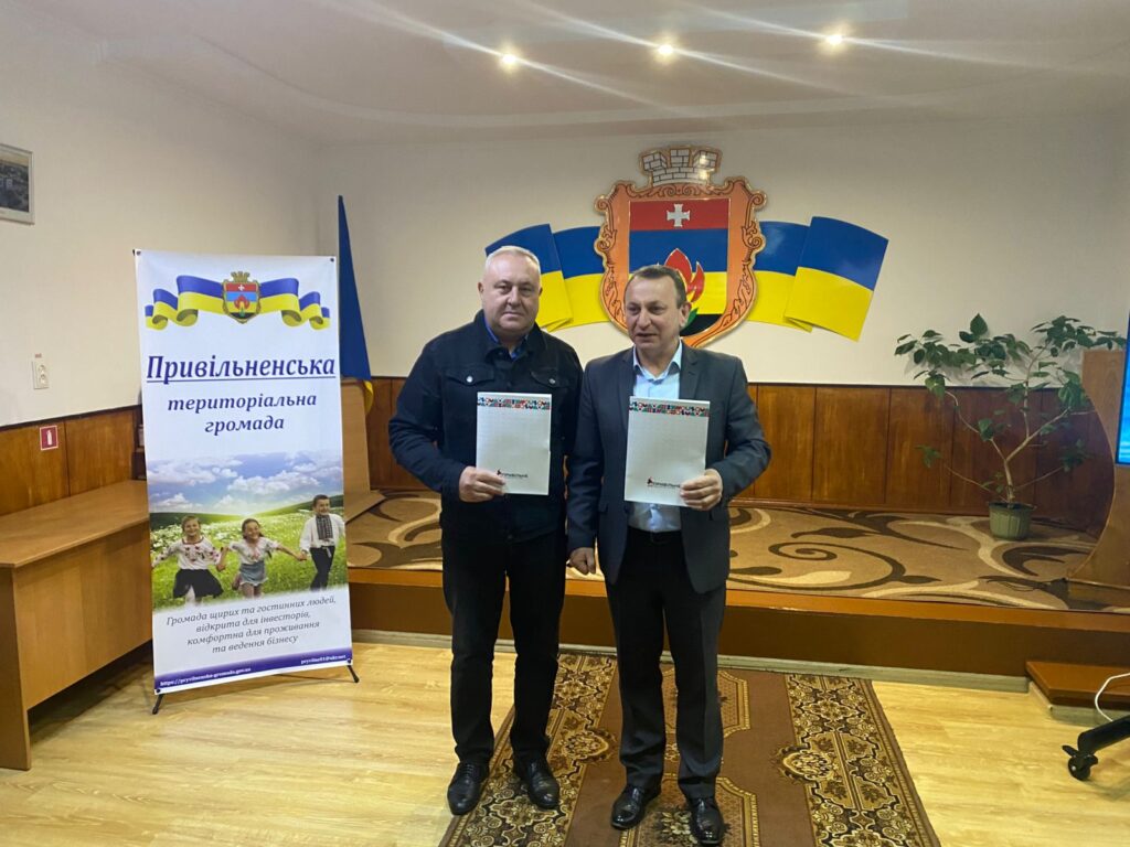 Двоє чоловіків стоять у формальному приміщенні, тримають документи. Позаду — емблема з щитом, хрестом і полум’ям, обрамлена українськими прапорами. Ліворуч — банер із написом «Привільненська територіальна громада» та фото дітей у полі. Обстановка — офіційна