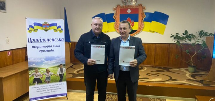 Двоє чоловіків стоять у формальному приміщенні, тримають документи. Позаду — емблема з щитом, хрестом і полум’ям, обрамлена українськими прапорами. Ліворуч — банер із написом «Привільненська територіальна громада» та фото дітей у полі. Обстановка — офіційна