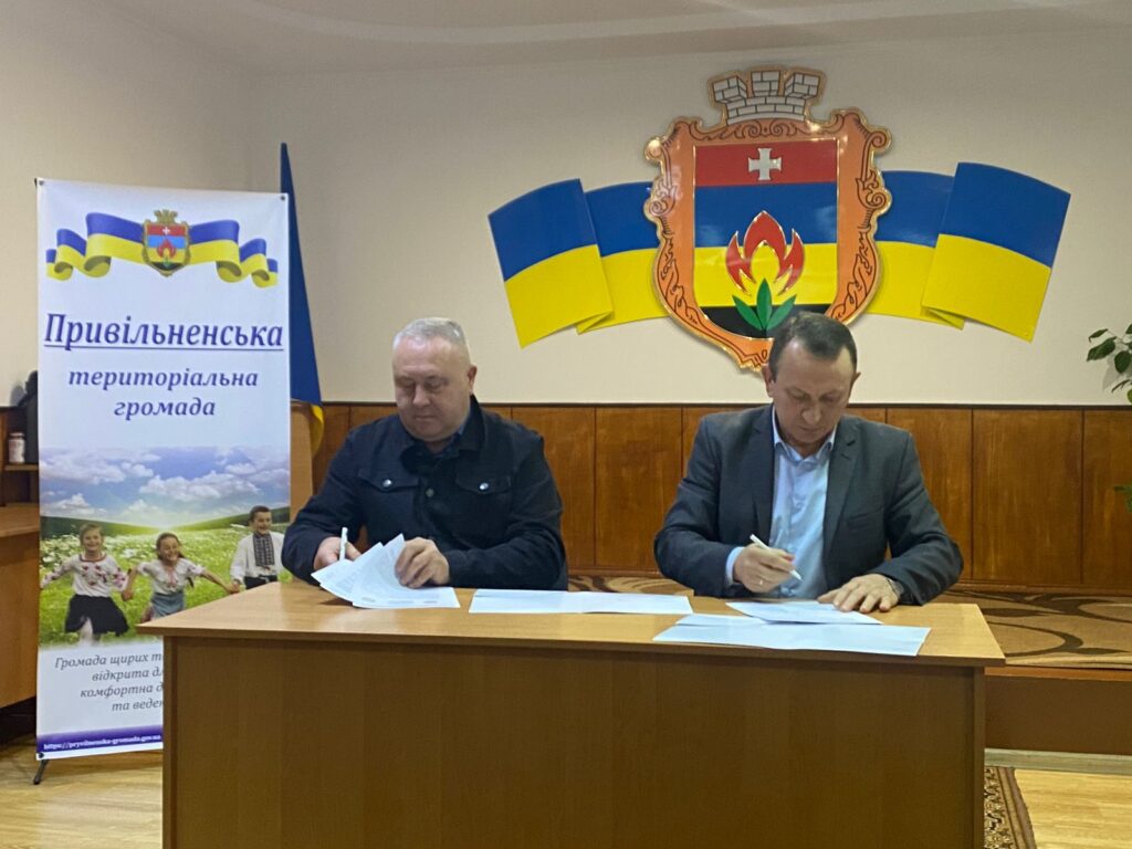 Двоє чоловіків сидять за дерев’яним столом і підписують документи. Позаду — емблема з червоним щитом, білим хрестом, стилізованим полум’ям і листком, обрамлена прапорами України. Ліворуч — банер із написом «Привільненська територіальна громада» та зображенням дітей у традиційному вбранні й пейзажем. Обстановка — офіційна