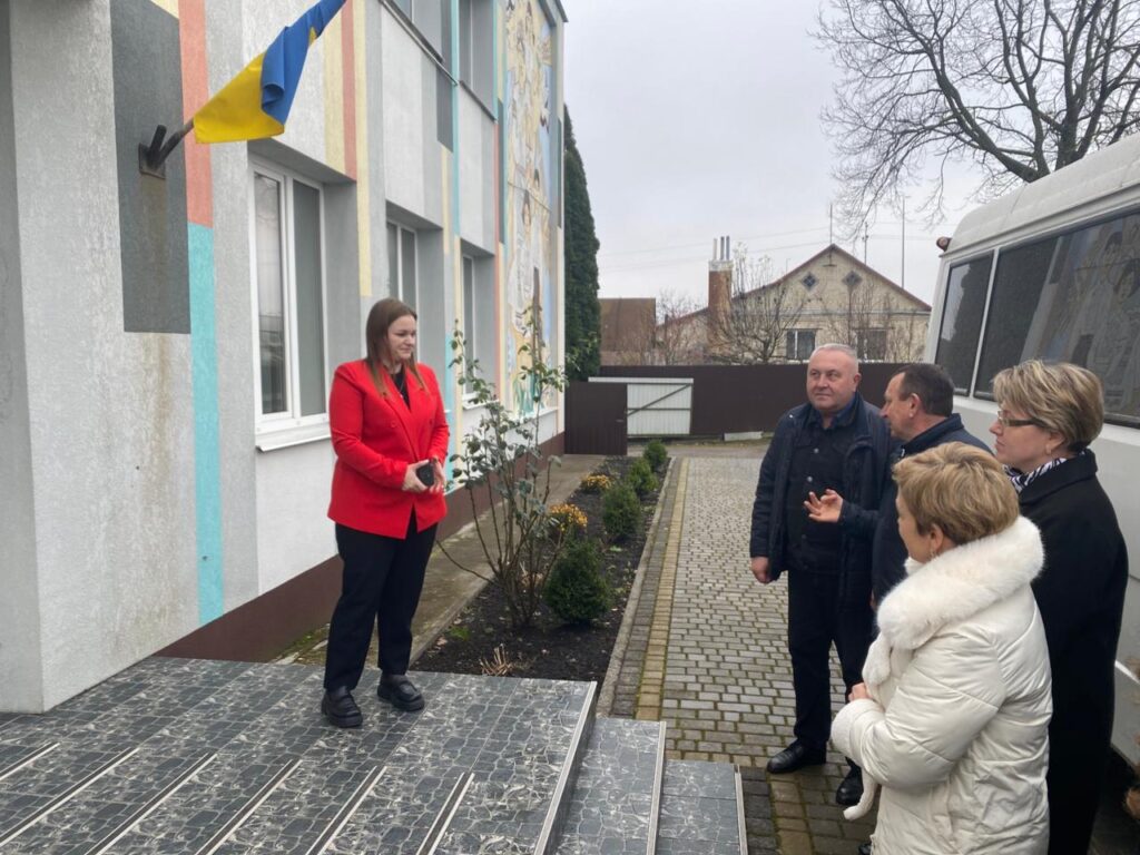 П’ятеро дорослих стоять на вулиці біля будівлі з яскравим фасадом. Жінка в червоному пальті стоїть на плитковій платформі біля входу, тримає телефон або пристрій. Четверо інших — на брукованій доріжці, звернені до неї. Біля входу — прапор України. На стіні — мурал, поруч — доглянутий садок і житлові будинки з голими деревами, що свідчить про холодну пору року.