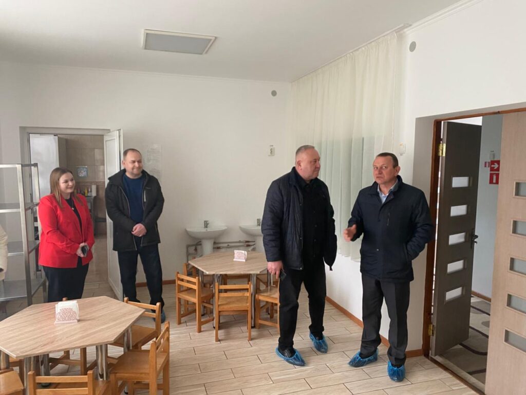 Четверо дорослих стоять у невеликій чистій кімнаті, схожій на дитячу їдальню або зону для занять. У приміщенні — маленькі дерев’яні стільці та шестикутні столи з тримачами для серветок. На стіні — дві раковини. Люди вдягнені в верхній одяг і мають на ногах сині бахіли, що свідчить про візит або інспекцію. Підлога вкрита плиткою, стіни білі, двері ведуть до іншого приміщення з плиткою