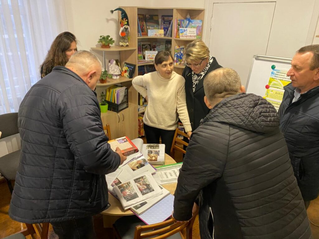 Шестеро дорослих зібрались навколо столу в кімнаті, схожій на бібліотеку або навчальний простір. На столі — відкриті книги, документи та друковані матеріали. Позаду — книжкові полиці з літературою, навчальними матеріалами та декором (рослина, лялька). Видно дошку з магнітами та плакатом. Люди обговорюють або вивчають матеріали. 