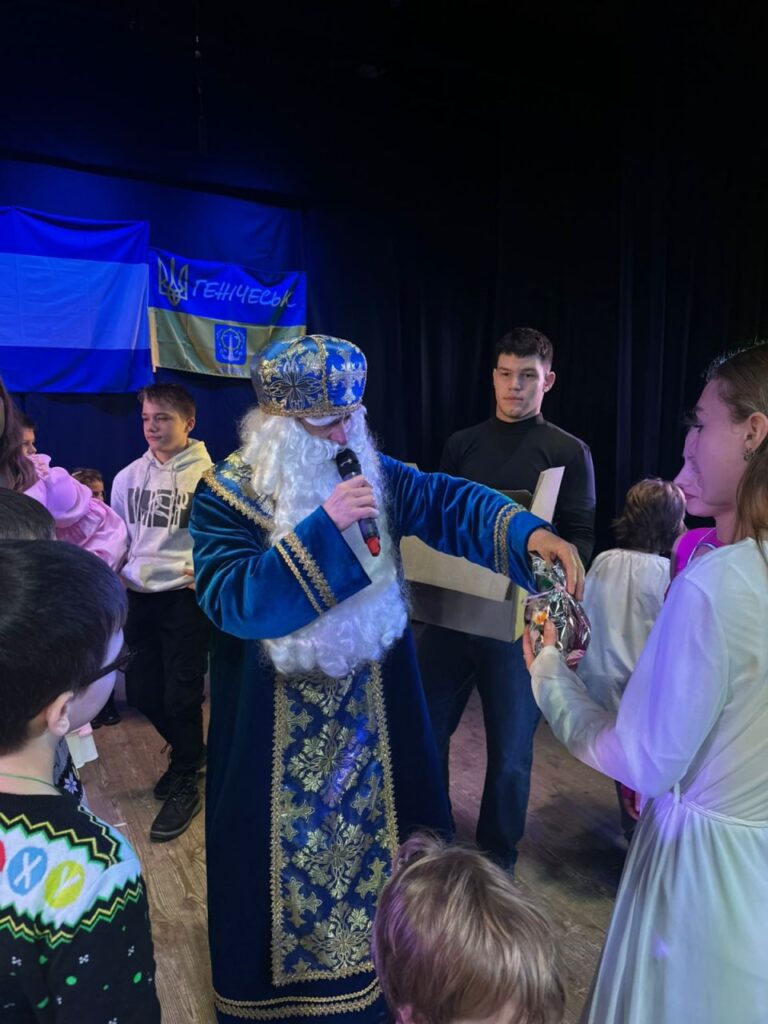 Людина в костюмі святого Миколая в синьо-золотій одежі з мікрофоном і подарунком спілкується з дітьми. Деякі діти й дорослі — у святковому вбранні.