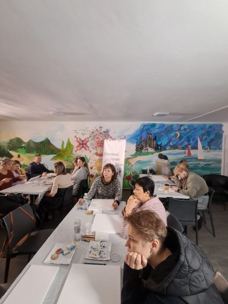 У приміщенні, схожому на клас або центр громади, дорослі й діти займаються малюванням. На столах — пензлі, кольорові картки, пляшки з водою. Жінка стоїть і допомагає учасникам. На стінах — мурали з природними сценами: гори, озеро з вітрильниками. Позаду — банер із написом «Слухаємо, Чуємо, Діємо». Атмосфера — тепла, творча, освітня.