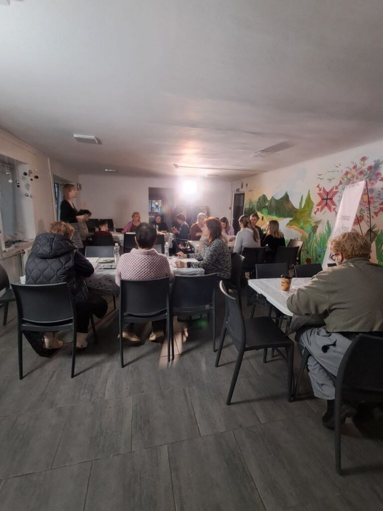 Група людей сидить за столами у світлій кімнаті, беручи участь у семінарі або тренінгу. Попереду — ведучий. На стіні — яскравий мурал із квітами, птахами та пагорбами. Видно фліпчарт із написом «Тренажер». Атмосфера — навчальна, творча, спільнотна.