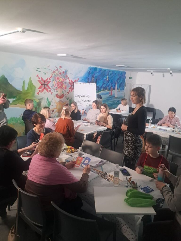 У кімнаті з муралом на стіні учасники сидять за столами з художніми матеріалами: пензлі, палітри, пляшки з водою, полотна. На муралі — пейзаж, місто, пара біля моря. У центрі — банер із написом «Слухаємо, Чуємо, Діємо». Атмосфера — творча, спільнотна, з елементами терапії або соціального залучення.