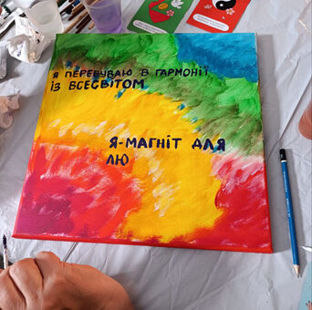 На столі — полотно з яскравим малюнком і написом українською: «Я перебуваю в гармонії із Всесвітом. Я — магніт для лю...». Поруч — пензлі, палітра, фіолетова рідина в чашці, ілюстративні картки. Видно руки людини, яка працює над картиною. Сцена — творча, з елементами самовираження та позитивних афірмацій.