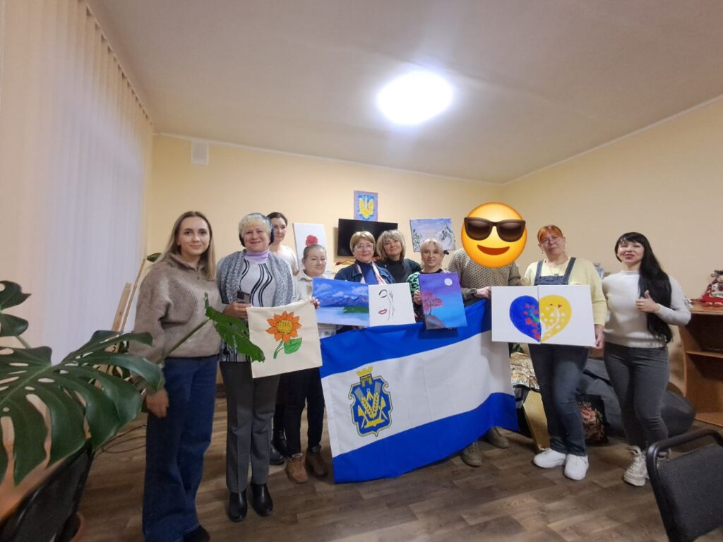 Десятеро людей стоять у кімнаті з бежевими стінами та дерев’яною підлогою, тримаючи власні художні роботи. Серед них — соняшник, абстракції, пейзаж, серце в синьо-жовтих кольорах із візерунками. У центрі — великий прапор із синьо-білим дизайном і гербом, схожим на тризуб. Позаду — телевізор і картини на стіні.