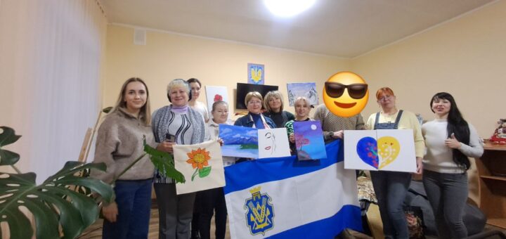 Десятеро людей стоять у кімнаті з бежевими стінами та дерев’яною підлогою, тримаючи власні художні роботи. Серед них — соняшник, абстракції, пейзаж, серце в синьо-жовтих кольорах із візерунками. У центрі — великий прапор із синьо-білим дизайном і гербом, схожим на тризуб. Позаду — телевізор і картини на стіні.