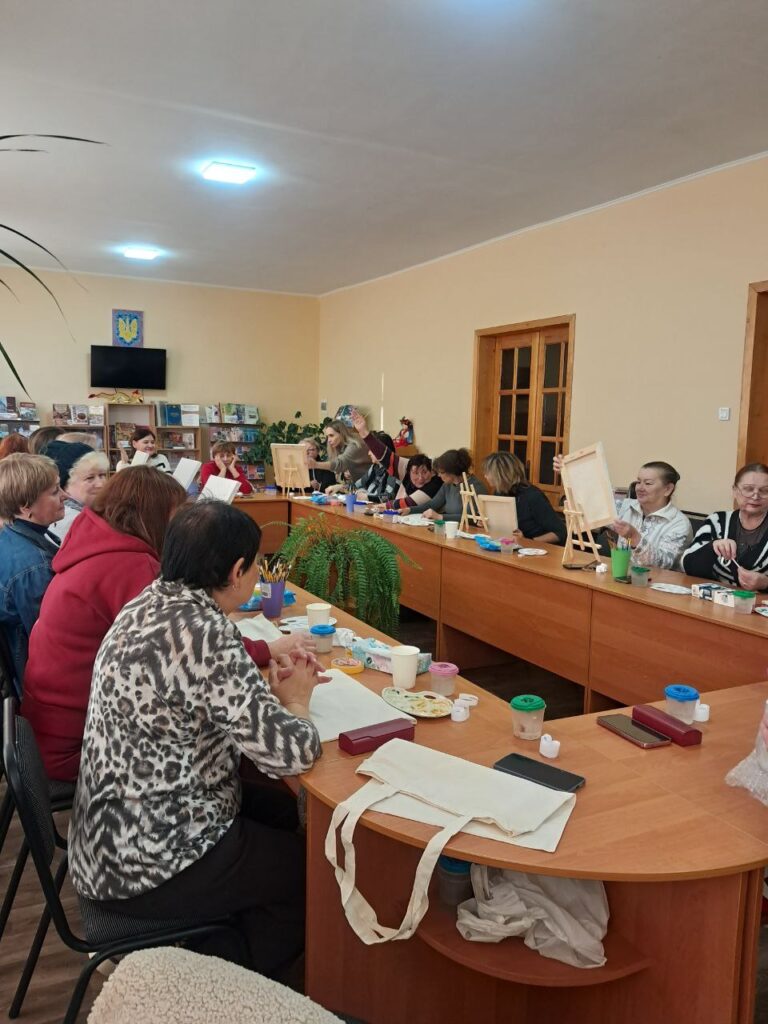 У кімнаті з дерев’яними столами учасники сидять у формі літери «П», займаються творчістю. На столах — мольберти, фарби, пензлі. Деякі малюють, інші спілкуються. Позаду — книжкові полиці, телевізор, емблема з тризубом, кімнатні рослини.