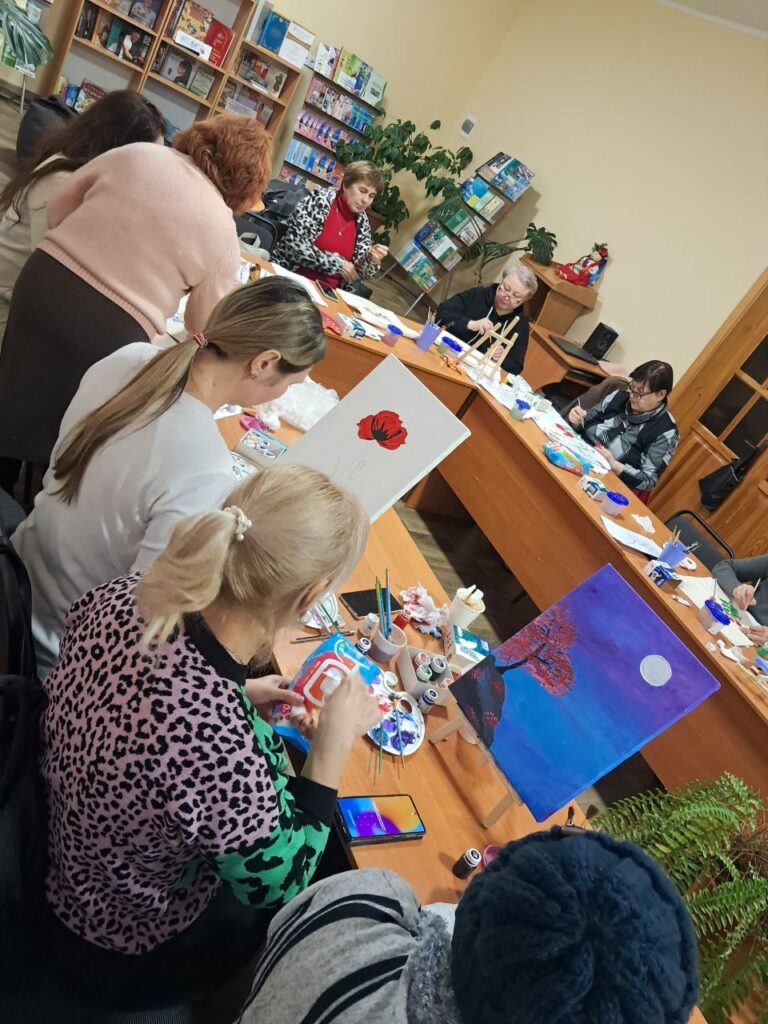 Група людей бере участь у художньому занятті в бібліотеці або центрі громади. Усі сидять за великим столом, малюють на полотнах. Видно червону квітку, пейзаж із деревом, пагорбом і місяцем на фіолетово-синьому небі. На стінах — книжкові полиці, кімнатні рослини.