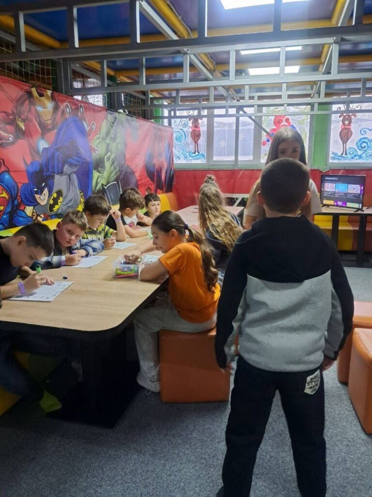 Група дітей сидить за прямокутним столом у яскравій кімнаті. Вони малюють або пишуть на аркушах, на столі — кольорові маркери й олівці. Стіни прикрашені зображеннями супергероїв: Бетмен, Супермен, Залізна людина, Халк. Стеля — з відкритими жовтими й фіолетовими трубами. На вікнах — святкові прикраси.