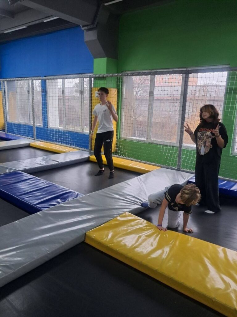 У критій батутній зоні — троє людей. Один стоїть на батуті, інший позує з жестом «peace», дитина повзає на м’якому маті. Підлога — з’єднані батутні секції, обрамлені кольоровими бортиками. Позаду — великі вікна з сіткою, стіни — зелені й сині.