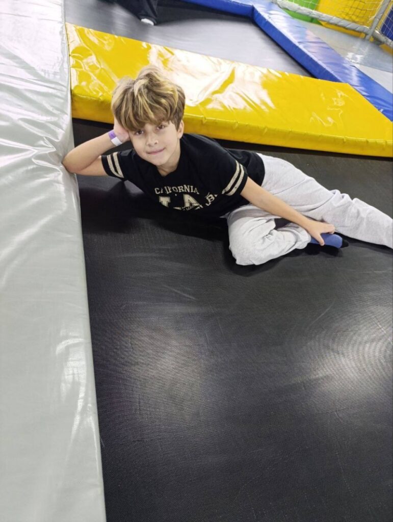 Дитина лежить на поверхні батута в критій зоні для активних ігор. Вона спирається на руку й дивиться в камеру. Одягнена в чорну футболку з написом «CALIFORNIA L.A.B.» та світло-сірі штани. Навколо — м’які кольорові борти: сірі, жовті, сині. Позаду — захисна сітка.