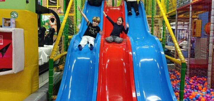 У критому ігровому просторі — чотири гірки: три сині з боків і одна червона посередині, що ведуть у басейн із кольоровими кульками (помаранчеві, фіолетові, сині, рожеві, білі). На вершині кожної гірки — дитина, готова спуститися. Конструкція оточена захисною сіткою, поруч — тунелі та зони для лазіння.