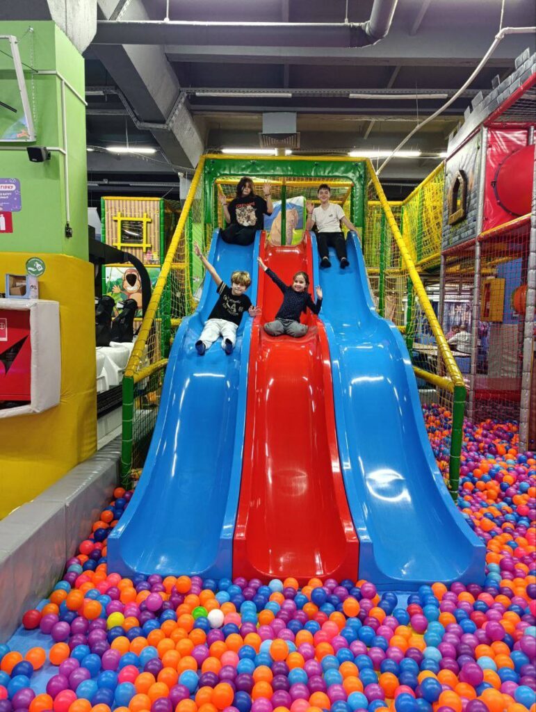 У критому ігровому просторі — чотири гірки: три сині з боків і одна червона посередині, що ведуть у басейн із кольоровими кульками (помаранчеві, фіолетові, сині, рожеві, білі). На вершині кожної гірки — дитина, готова спуститися. Конструкція оточена захисною сіткою, поруч — тунелі та зони для лазіння. 