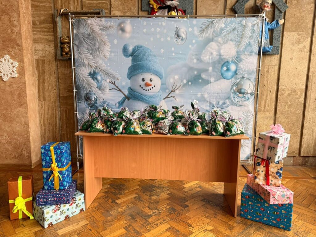 У центрі — дерев’яний стіл із численними зеленими подарунковими пакетами в блискучій обгортці. Позаду — великий банер із усміхненим сніговиком у синій шапці та шарфі, серед засніжених гілок і синіх прикрас. По боках — стопки подарункових коробок у яскравому пакуванні.