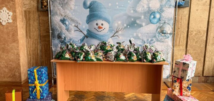 У центрі — дерев’яний стіл із численними зеленими подарунковими пакетами в блискучій обгортці. Позаду — великий банер із усміхненим сніговиком у синій шапці та шарфі, серед засніжених гілок і синіх прикрас. По боках — стопки подарункових коробок у яскравому пакуванні.