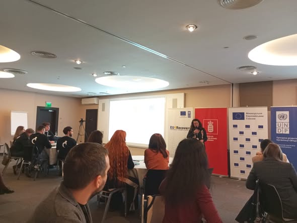 Конференц-зал із учасниками за круглими столами. Спікер виступає перед екраном, поруч — три вертикальні банери: «EU4Recovery», емблема з червоним фоном, логотип ПРООН із кириличним текстом. Освітлення — яскраве, атмосфера — професійна, міжнародна. Сцена — презентація або семінар за участі ЄС і ПРООН.