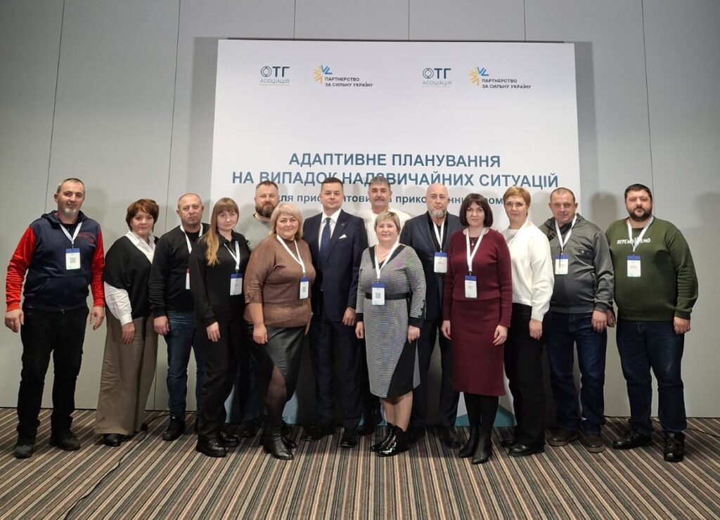 Тринадцять людей стоять перед банером із логотипами «ОТГ Асоціація» та «Партнерство за сильну Україну». Тема — адаптивне планування для прикордонних громад. Усі учасники — з бейджами, одяг — діловий.