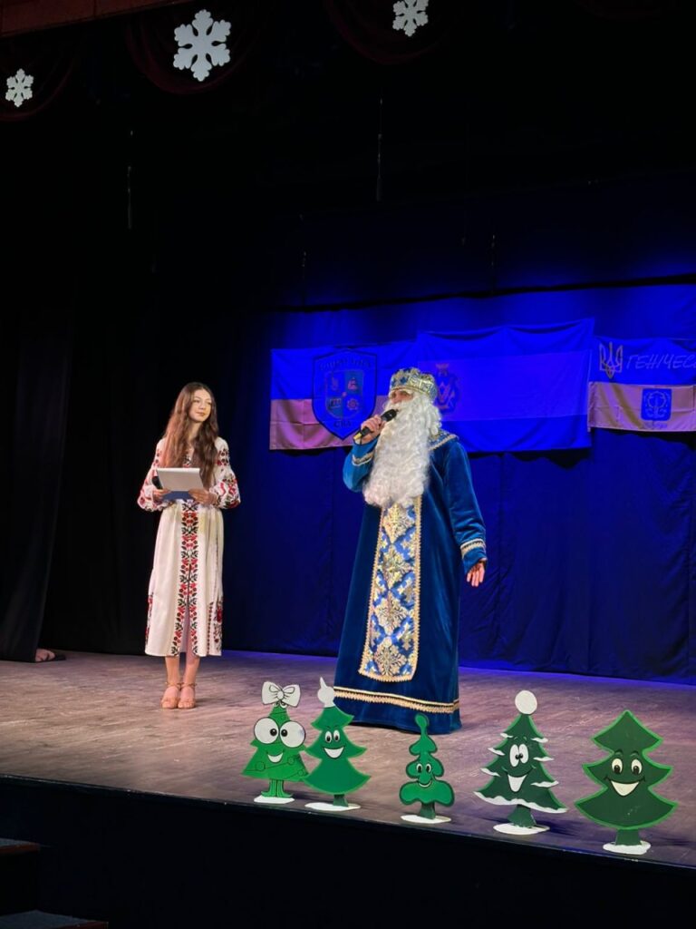 На сцені двоє людей: один — у вишиванці з планшетом, інший — у костюмі святого Миколая з білою бородою, синьо-золотим халатом і короною. Вони виступають із мікрофонами. Декор — мультяшні ялинки, сніжинки, прапори з емблемами.