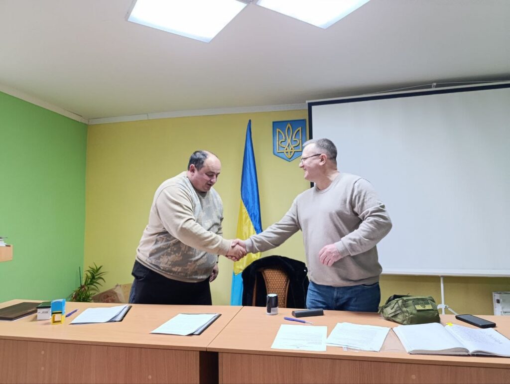 Два чоловіка у світлому кабінеті тиснуть один одному руки. Перед ними на столах лежать документи, позаду - прапор України.