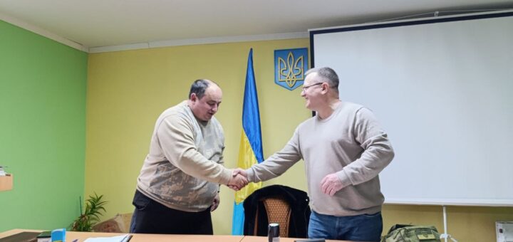 Два чоловіка у світлому кабінеті тиснуть один одному руки. Перед ними на столах лежать документи, позаду - прапор України.