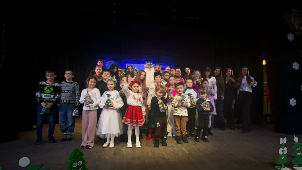 На сцені група дітей і дорослих позує для фото. Діти — у святковому та традиційному одязі, деякі — з віночками. Багато хто тримає подарункові пакети. Декор — темна завіса, синє освітлення, мультяшні ялинки та сніжинки.