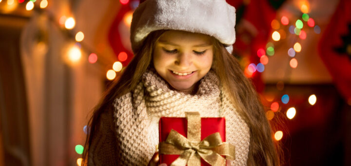 Дитина в червоному ковпаку Санти та товстому в’язаному шарфі тримає червону подарункову коробку з золотою стрічкою. Позаду — яскраві новорічні вогники та святкові декорації.