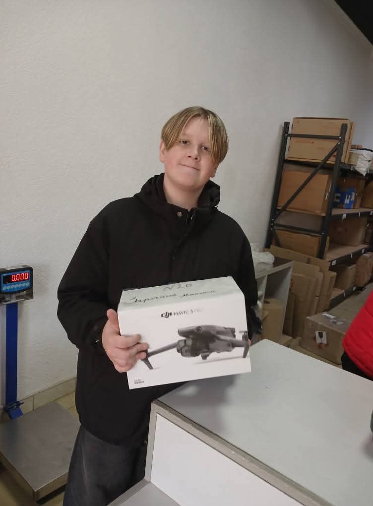 У приміщенні стоїть людина з коробкою дрона DJI Mavic 3. На коробці — зображення дрона та напис «DJI MAVIC 3». Зверху — рукописний напис: «N20» і «Зручний дрончик». Позаду — полиці з картонними коробками та пакувальними матеріалами, що нагадує склад або зону зберігання.