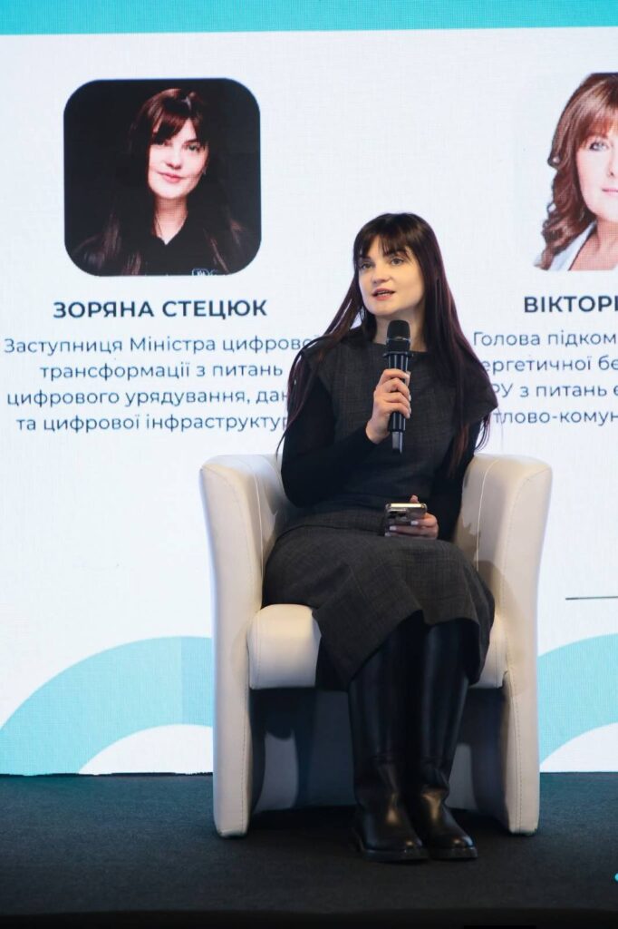 Жінка сидить на білому кріслі, тримає мікрофон і смартфон, ймовірно, бере участь у панельній дискусії. Позаду — великий екран із двома портретами та текстом українською. Один із написів: «Зоряна Стецюк — заступниця Міністра цифрової трансформації з питань цифрового урядування, даних та цифрової інфраструктури».