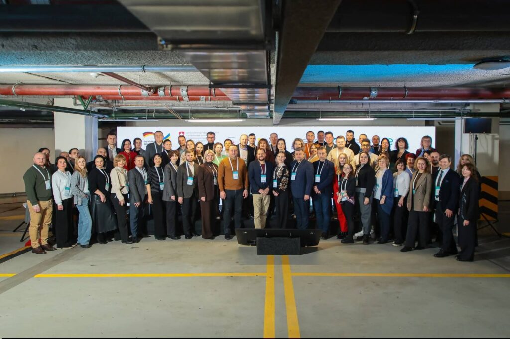 Велика група людей у діловому одязі позує для фото в критому паркінгу. Позаду — білий банер із логотипами, зокрема «german cooperation» та «Swiss Confederation». Усі учасники мають бейджі, що свідчить про участь у конференції або семінарі.