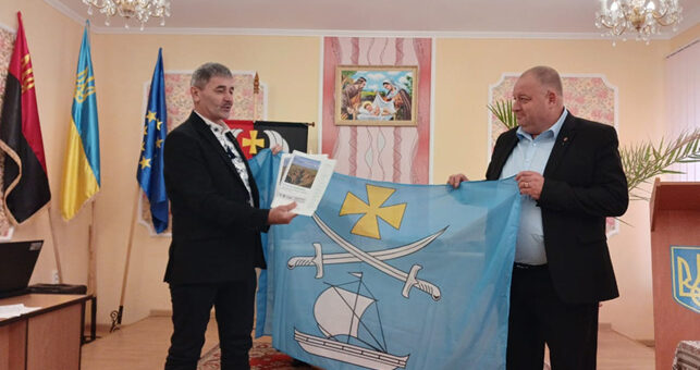 Двоє чоловіків стоять у формальному приміщенні, тримають світло-блакитний прапор із жовтим хрестом, двома шаблями та білим вітрильником. Один із них також тримає документ або буклет із фотографією на обкладинці. Позаду — кілька прапорів, зокрема український і прапор ЄС. У кімнаті — люстра, релігійна картина на стіні та трибуна з тризубом.