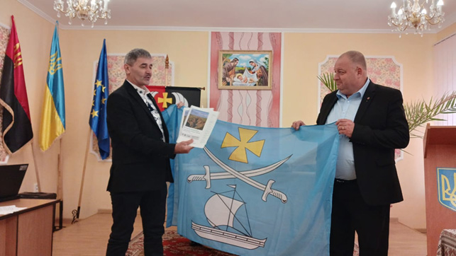 Двоє чоловіків стоять у формальному приміщенні, тримають світло-блакитний прапор із жовтим хрестом, двома шаблями та білим вітрильником. Один із них також тримає документ або буклет із фотографією на обкладинці. Позаду — кілька прапорів, зокрема український і прапор ЄС. У кімнаті — люстра, релігійна картина на стіні та трибуна з тризубом.