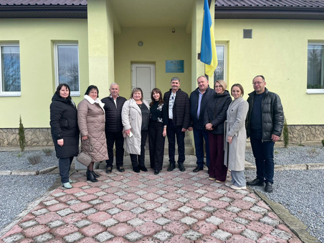 Десятеро дорослих стоять перед світло-жовтою будівлею з черепичним дахом і кам’яним цоколем. Група розташована на декоративній кам’яній доріжці біля входу. Над дверима — синьо-жовтий прапор України та синя табличка з текстом. Люди вдягнені в зимовий одяг.