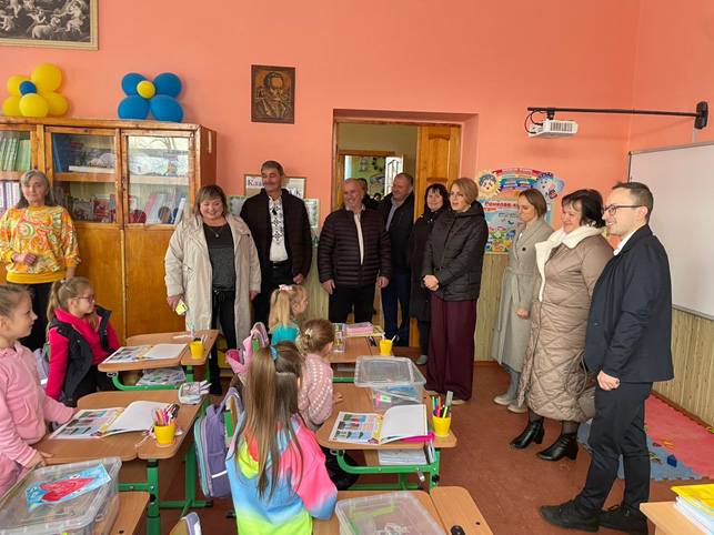 У класі сидять діти за партами, зайняті завданням. На столах — кольорові матеріали, книжки, жовті стаканчики з ручками та прозорі контейнерчики. Попереду — група дорослих у формальному або напівформальному одязі, які спостерігають або взаємодіють із дітьми. Стіни — теплого персикового кольору, є дошка, навчальні плакати, шафа з книжками, повітряні кульки та релігійне зображення. 
