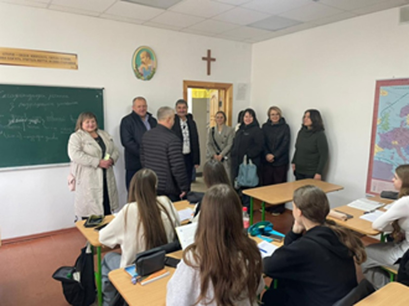 У класі учні сидять за партами, працюють із зошитами та письмовими матеріалами. Попереду — група дорослих, ймовірно, працівники школи або відвідувачі. На стіні — карта Європи, релігійні символи (хрест і зображення святого), а також дошка з написом українською, де згадується «експоненціальне рівняння», «логарифм» і «приклад».