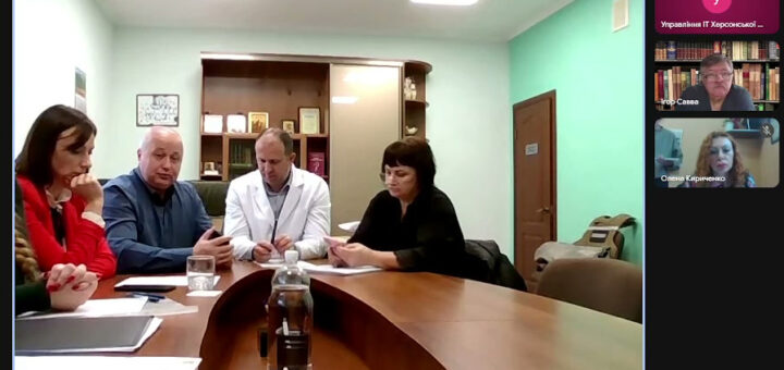 У головному вікні відеоконференції — п’ятеро людей за вигнутим столом у світлій офісній кімнаті. Один із них у білому халаті. На столі — документи, блокноти, склянки з водою. Позаду — шафа з книгами, нагородами та рамками. Праворуч — три менші відеовікна: «Управління ІТ Херсонської ...», «Ігор Савва» та «Олена Кириченко». Внизу — панель керування, час — 12:07.