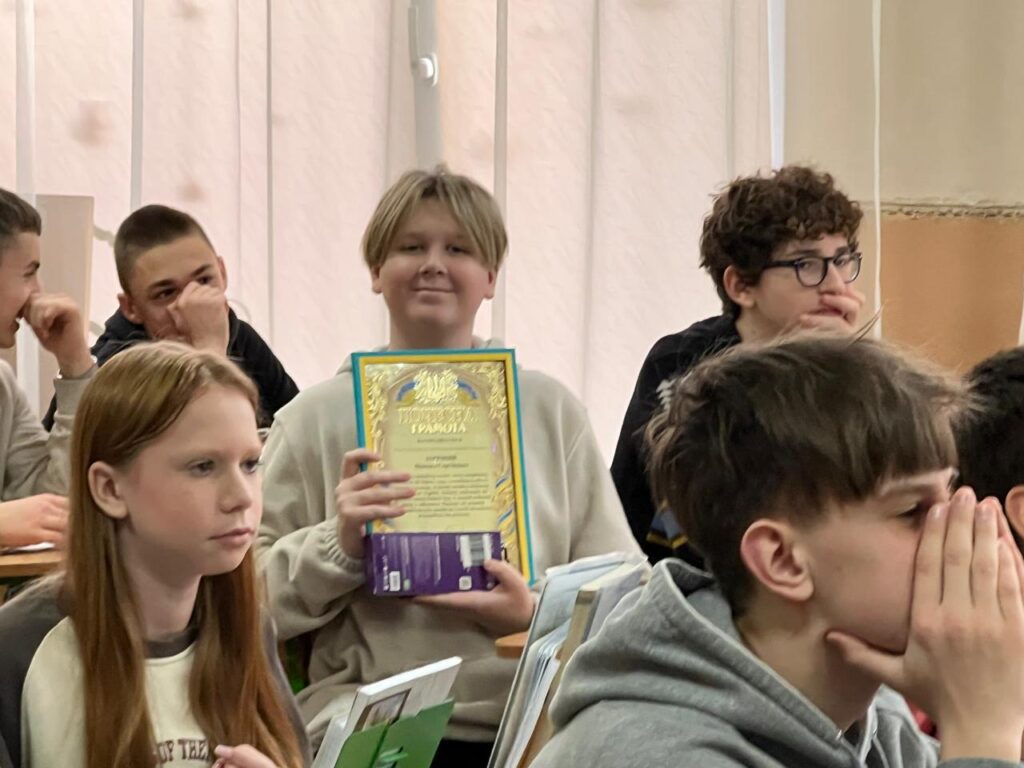 Максим Зарічний тримає Почесну грамоту з декоративною рамкою, демонструючи її присутнім.