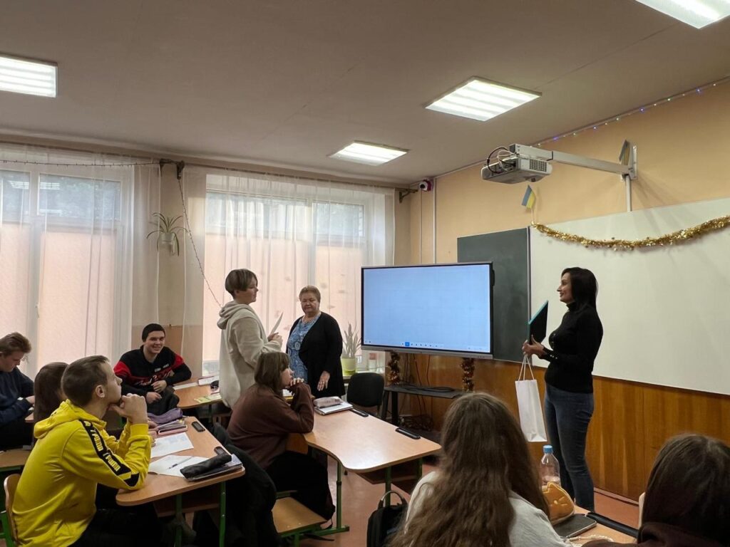 Максим Зарічний стоїть попереду, тримає Почесну грамоту та подарунок. Поруч — Тетяна Гасаненко, яка вітає його. У залі — люди, святкові прикраси, український прапор, біла дошка та цифровий екран.