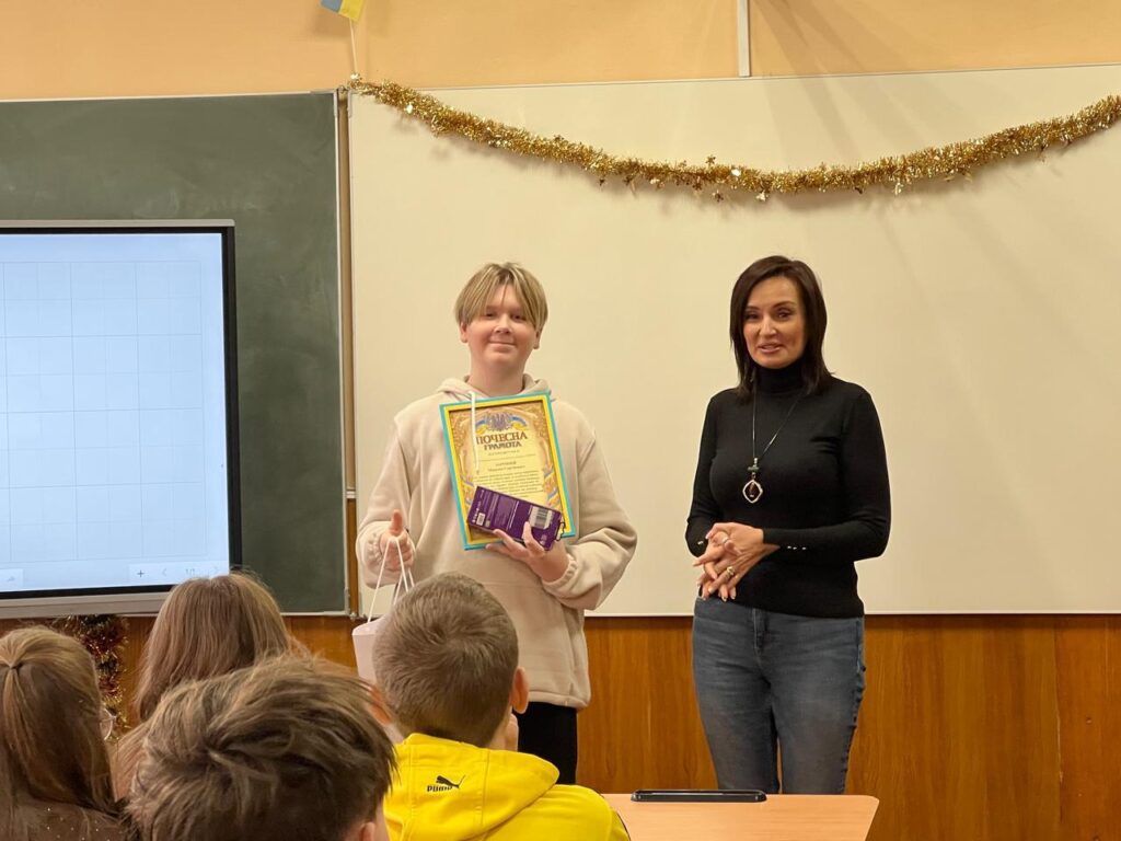 Максим Зарічний стоїть попереду, тримає Почесну грамоту та подарунок. Поруч — Тетяна Гасаненко, яка вітає його. У залі — люди, святкові прикраси, український прапор, біла дошка та цифровий екран.