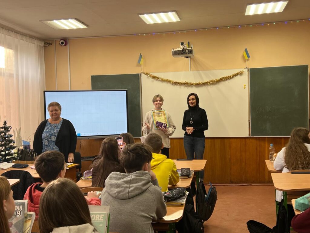 У залі проходить урочиста подія. Максим Зарічний, 13-річний волонтер, стоїть попереду, поруч із Тетяною Гасаненко. На фоні — біла дошка, цифровий екран, новорічна гірлянда та прапори України.