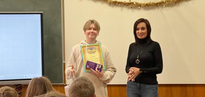 Максим Зарічний стоїть попереду, тримає Почесну грамоту та подарунок. Поруч — Тетяна Гасаненко, яка вітає його. У залі — люди, святкові прикраси, український прапор, біла дошка та цифровий екран.