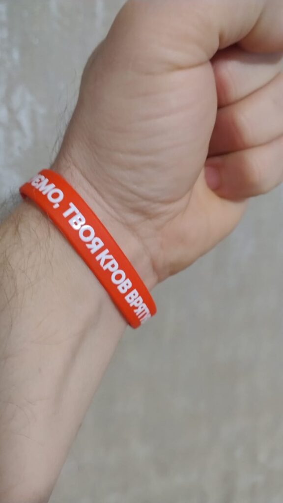 Крупний план зап’ястя людини з червоним силіконовим браслетом. На браслеті — білий напис кирилицею: «Твоя кров врятує життя».