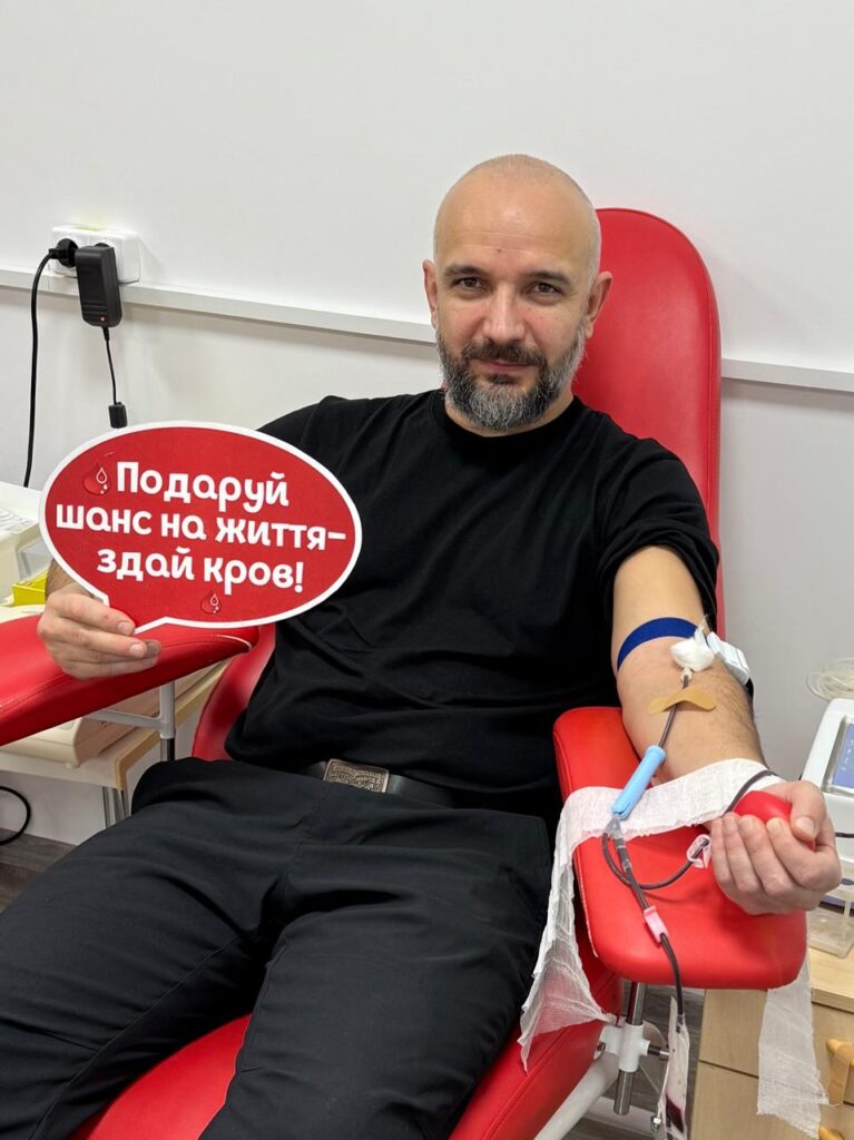 Юліан Лучко  сидить у червоному медичному кріслі, здає кров. У руці — червона табличка у формі хмаринки з білим написом українською: «Подаруй шанс на життя — здай кров!». У руку вставлено голку, під’єднану до трубки та медичного обладнання. 