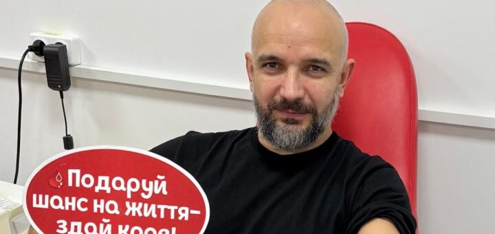 Юліан Лучко сидить у червоному медичному кріслі, здає кров. У руці — червона табличка у формі хмаринки з білим написом українською: «Подаруй шанс на життя — здай кров!». У руку вставлено голку, під’єднану до трубки та медичного обладнання.