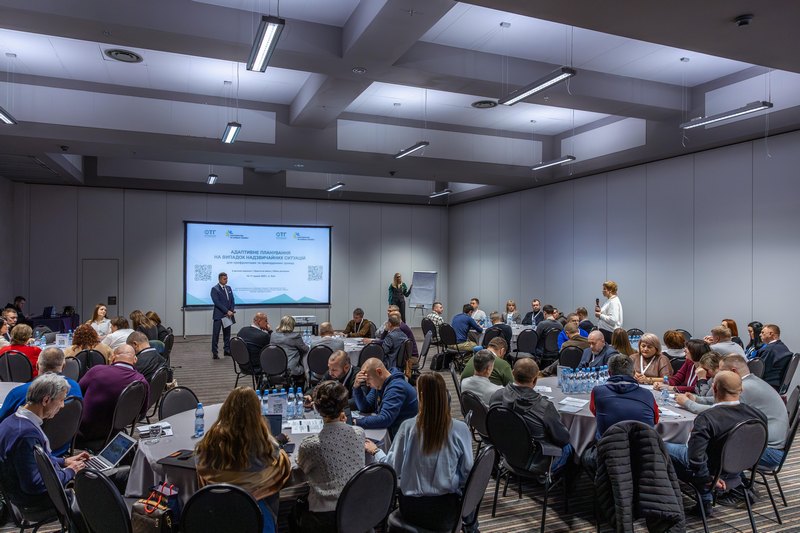 Світла конференц-зала з круглими столами, за якими сидять учасники. На передньому плані — екран із презентацією російською мовою: «Адаптивные подходы на выборах представительных органов». Один спікер виступає біля екрана, інший — праворуч у залі. На столах — пляшки з водою, документи.